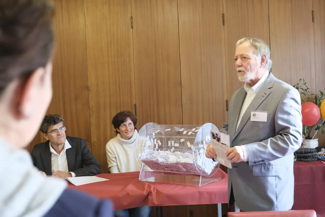 Der Vorstandsvorsitzende steht neben einer durchsichtigen Lotterietrommel voller Lose und spricht; zwei weitere Personen sitzen am Tisch.